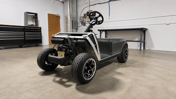 A custom-built, stripped-down electric golf cart with a black body, white side panels, and a steering wheel, parked inside a clean workshop with concrete floors and toolboxes.