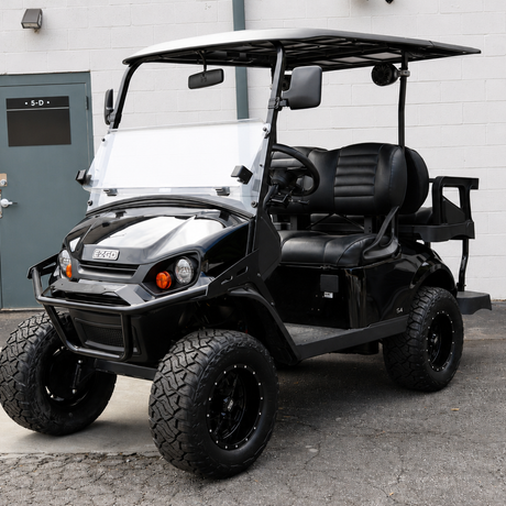 A black and white custom-built electric golf cart with off-road tires, a white canopy roof, and a custom front bumper, parked in front of a white brick building.
