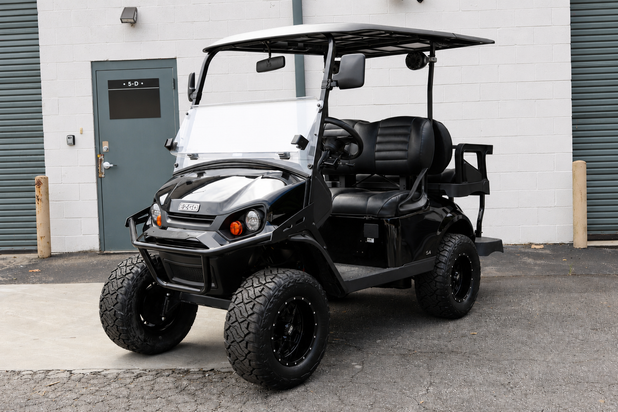 A black and white custom-built electric golf cart with off-road tires, a white canopy roof, and a custom front bumper, parked in front of a white brick building.