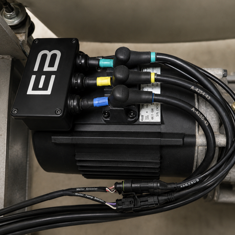 A close-up of an electric motor assembly with a black control box labeled EB, multiple black cables with colored connectors, and a motor encoder cable, photographed in an industrial setting.