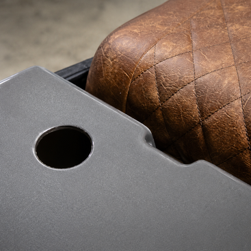Close-up of a gray metal tray table with a circular cutout, positioned next to a brown quilted leather seat.